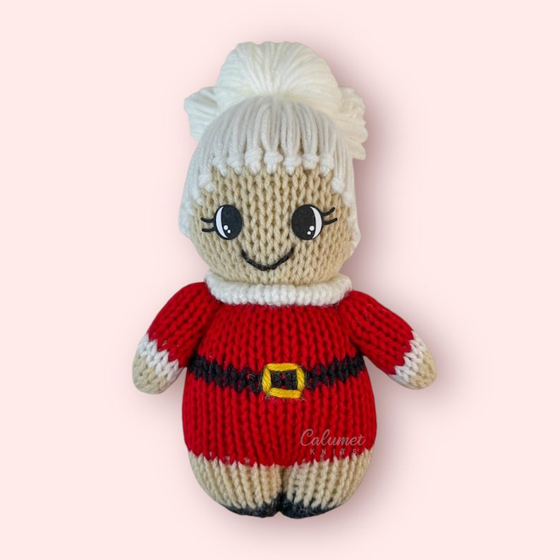K&ouml;nnte beinhalten: Gestrickte Weihnachtspuppe mit einem rot-wei&szlig;en Weihnachtsmannkost&uuml;m, wei&szlig;en Haaren und einem schwarzen G&uuml;rtel mit goldener Schnalle. Die Puppe hat ein l&auml;chelndes Gesicht mit schwarzen Augen und einem wei&szlig;en Kragen.