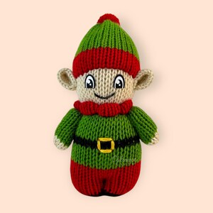 K&ouml;nnte beinhalten: Eine gestrickte Elfenpuppe mit gr&uuml;nem Hut, rot-gr&uuml;nem gestreiftem K&ouml;rper und einem schwarzen G&uuml;rtel. Der Elf hat ein l&auml;chelndes Gesicht mit schwarzen Augen und einem roten Mund.
