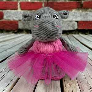 May include: A gray knitted hippopotamus wearing a pink tutu. The hippopotamus has large black eyes and a pink nose. The tutu is made of pink tulle.