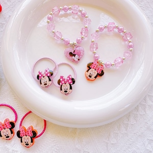 May include: Pink beaded bracelets and hair ties featuring Minnie Mouse. The bracelets have charms with Minnie Mouse and heart-shaped pendants. The hair ties have Minnie Mouse faces with pink bows. The items are arranged on a white plate.