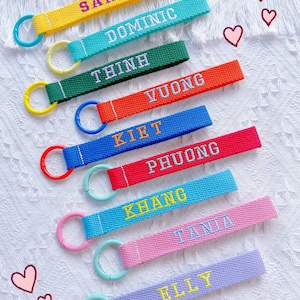 May include: A selection of brightly coloured keyrings, each embroidered with a name. The keyrings come in various colours such as yellow, teal, green, red, blue, and pink. The names are embroidered in contrasting colours. Each keyring has a matching ring.