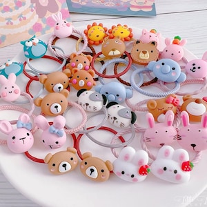 May include: A collection of colorful hair ties with animal-themed designs, including bears, bunnies, lions, and owls. The elastic bands are in various colors, and the decorative elements are made of a soft, textured material. The hair accessories are arranged on a white plate.