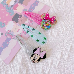 Op de afbeelding: Drie kleurrijke haarclips. Eén roze glitterclip met een Minnie Mouse strik, één lichtblauwe clip met groene sterren en één zilveren glitterclip met een Minnie Mouse gezicht. De clips liggen op een roze achtergrond met patronen.