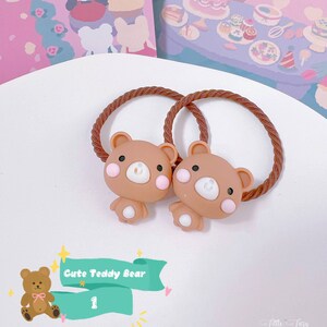 May include: Two brown teddy bear hair ties with pink cheeks and white accents. The hair ties have a brown rope-like texture. The image also includes a teal banner with the text "Cute Teddy Bear" and the number "1".
