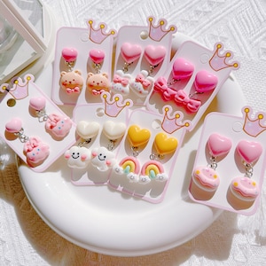 以下が含まれることがあります： ハート、リボン、クマ、雲、虹、ケーキなどのデザインのクリップオンイヤリングのコレクション。 イヤリングは主にピンク、白、黄色で、王冠のアクセントが付いたピンクのカードに表示されています。