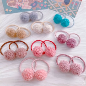 May include: A selection of pom-pom hair ties in shades of pink, red, brown, blue, and beige. Each tie has a pair of fluffy pom-poms attached to a matching elastic band. The background is a soft, white surface.