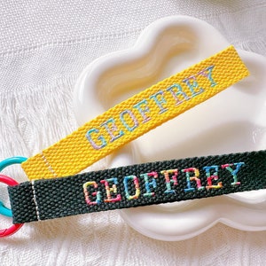 Könnte beinhalten: Zwei personalisierte Schlüsselanhänger mit dem Namen "GEOFFREY" in bunten Fäden gestickt. Einer ist gelb, der andere dunkelgrün. Jeder hat einen unterschiedlich farbigen Karabiner: rot und blaugrün. Die Schlüsselanhänger liegen auf einer weißen, wolkenförmigen Schale.