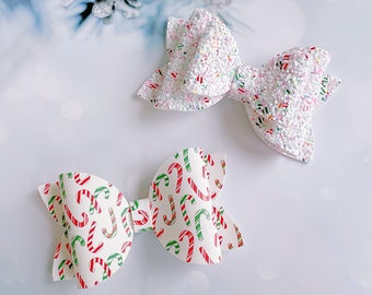 May include: Two Christmas-themed hair bows. One bow is white with a glittery finish and colorful sprinkles. The other bow is white with a repeating pattern of red and green candy canes. The bows are on a white background.