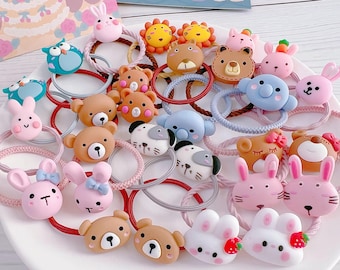 May include: A collection of colorful hair ties with animal-themed designs, including bears, bunnies, lions, and owls. The elastic bands are in various colors, and the decorative elements are made of a soft, textured material. The hair accessories are arranged on a white plate.