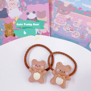 May include: Two brown hair ties with a cute teddy bear design. Each bear has a bow tie and a heart on its cheek. The background features illustrations of teddy bears and the text "Cute Teddy Bear".