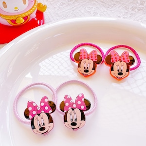 May include: Four Minnie Mouse hair ties with pink bows and a pink elastic band. The Minnie Mouse faces are brown, with a pink bow with white polka dots. The hair ties are on a white plate.