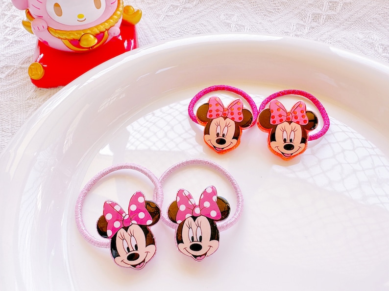 May include: Four Minnie Mouse hair ties with pink bows and a pink elastic band. The Minnie Mouse faces are brown, with a pink bow with white polka dots. The hair ties are on a white plate.