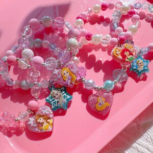 Könnte beinhalten: Rosa und weiße Armbänder mit Anhängern zum Thema Prinzessin. Die Armbänder haben verschiedene Perlen Größen und Formen, darunter klare, glitzernde und perlenartige Perlen. Zu den Anhängern gehören Herz- und Sternformen mit Prinzessinnenfiguren.
