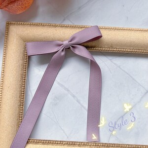 May include: A mauve ribbon bow with long tails sits on a gold picture frame. The text "Style 3" is written in a cursive font with small butterfly illustrations. The background is a marble-effect surface.