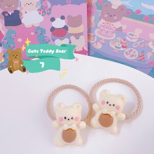 May include: Two cream-colored teddy bear hair ties with brown accents and pink cheeks. The bears are attached to beige elastic bands. The background features illustrations of teddy bears and the text "Cute Teddy Bear".