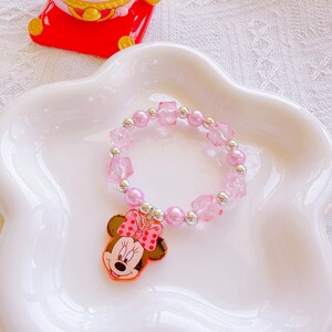 May include: A pink beaded bracelet with a Minnie Mouse charm. The bracelet features pink beads, clear square beads, and silver accents. The Minnie Mouse charm has a red bow with white polka dots.