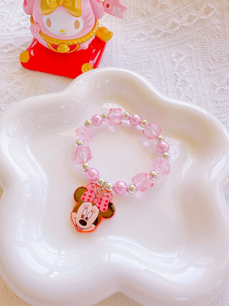 May include: A pink beaded bracelet with a Minnie Mouse charm. The bracelet features pink beads, clear square beads, and silver accents. The Minnie Mouse charm has a red bow with white polka dots.