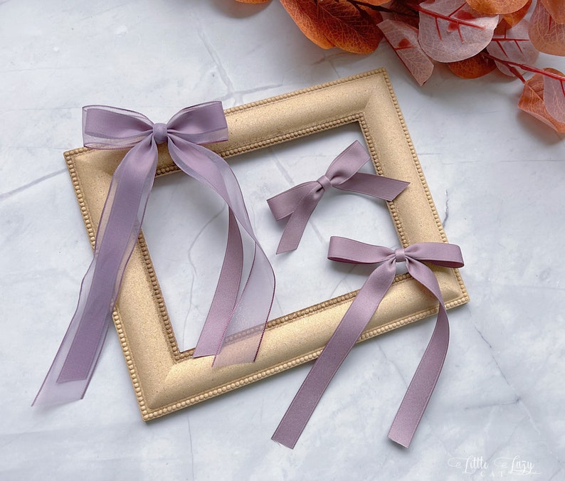 May include: Three lavender hair bows of different sizes. One bow features long, sheer ribbons. The bows are arranged on a gold picture frame. The background has a marble effect.