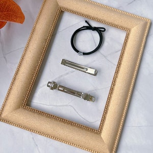 May include: A selection of hair accessories, comprising two silver barrettes and a black elastic hair tie. These items are arranged within a light brown picture frame. The frame features a textured surface and a beaded border.