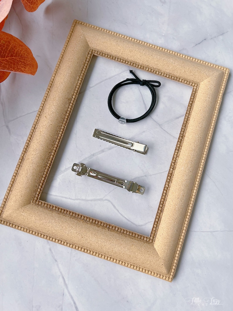 May include: A selection of hair accessories, comprising two silver barrettes and a black elastic hair tie. These items are arranged within a light brown picture frame. The frame features a textured surface and a beaded border.