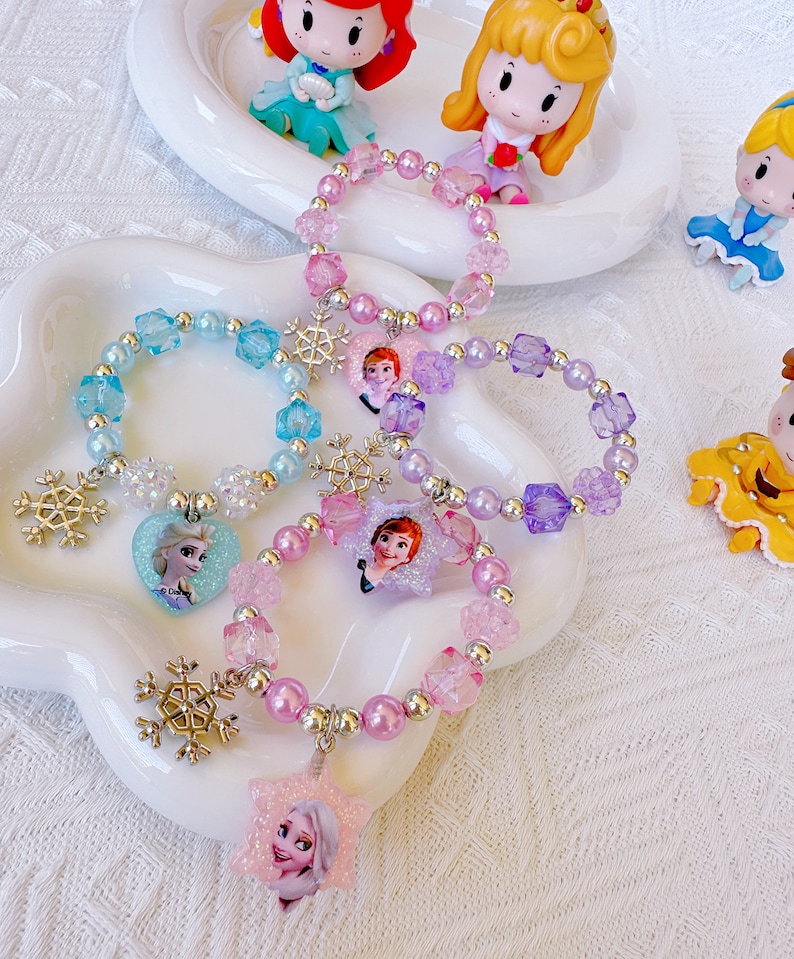 K&ouml;nnte beinhalten: Eine Sammlung von Armb&auml;ndern mit Perlen in Rosa, Lila und Blau, verziert mit Schneeflocken-Anh&auml;ngern und Charakter-Anh&auml;ngern. Die Armb&auml;nder haben Perlen und Kristallperlen mit Bildern von Zeichentrickfiguren.