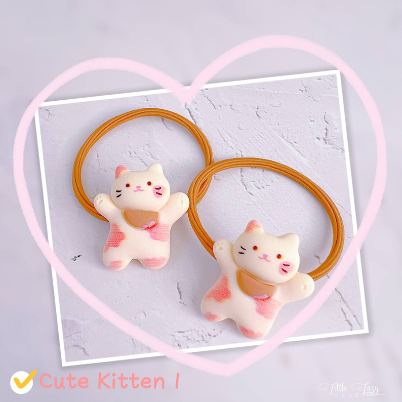 Puede incluir: Dos gomas para el pelo con un dise&ntilde;o de gatito. Los gatitos son blancos con manchas rosas y un babero marr&oacute;n claro. Las gomas para el pelo son de color marr&oacute;n claro. El texto "Cute Kitten I" est&aacute; en la esquina inferior izquierda.