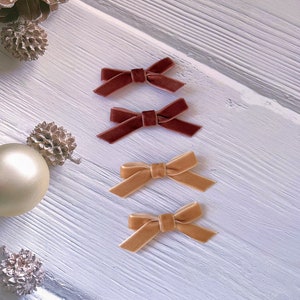 May include: Four velvet bows in shades of brown and gold are arranged on a white, textured surface. Two bows are a deep burgundy, and two are a light gold color. Decorative pinecones and a gold ornament are also present.