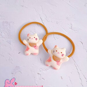 Puede incluir: Dos gomas para el pelo con un dise&ntilde;o de gatito. Los gatitos son blancos con manchas rosas y un babero rosa. Las gomas para el pelo son de color marr&oacute;n claro. El texto "Cute Kitten 1" es visible.