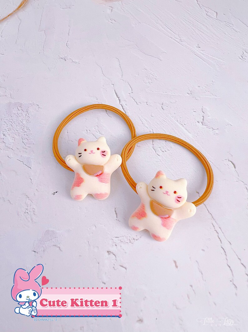 Puede incluir: Dos gomas para el pelo con un dise&ntilde;o de gatito. Los gatitos son blancos con manchas rosas y un babero rosa. Las gomas para el pelo son de color marr&oacute;n claro. El texto "Cute Kitten 1" es visible.