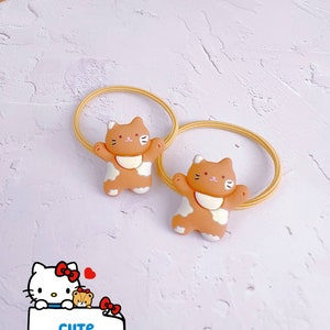 Puede incluir: Dos coleteros con bandas el&aacute;sticas color beige y dijes con forma de gato. Los dijes son de color beige y blanco con detalles negros. La imagen tambi&eacute;n incluye un gr&aacute;fico de Hello Kitty con el texto "CUTE KITTEN 2".