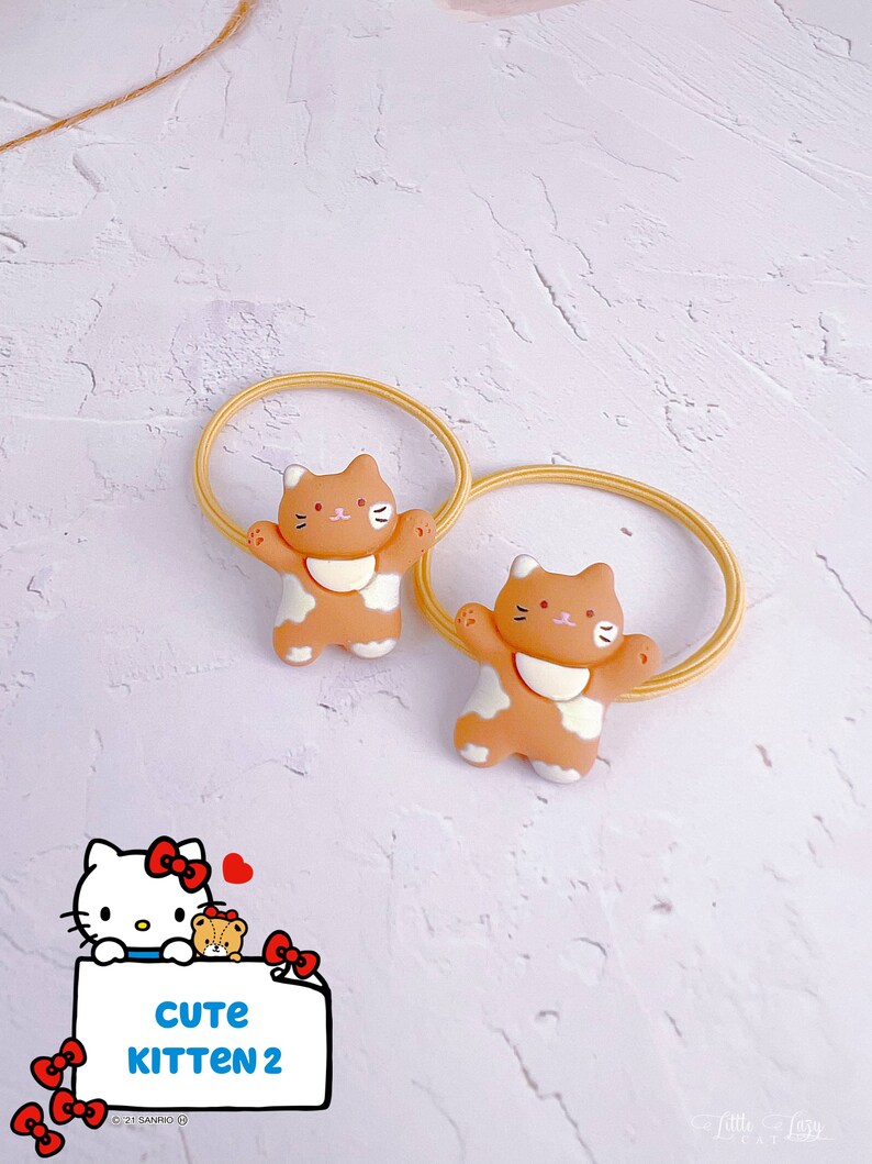 Puede incluir: Dos coleteros con bandas el&aacute;sticas color beige y dijes con forma de gato. Los dijes son de color beige y blanco con detalles negros. La imagen tambi&eacute;n incluye un gr&aacute;fico de Hello Kitty con el texto "CUTE KITTEN 2".