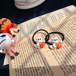 Pode incluir: Duas presilhas de cabelo com tema de Halloween com fantasmas brancos segurando ab&oacute;boras laranja. As faixas el&aacute;sticas pretas est&atilde;o em um fundo de papel vintage, com figuras de ab&oacute;bora e esqueleto. O tema &eacute; o Halloween.