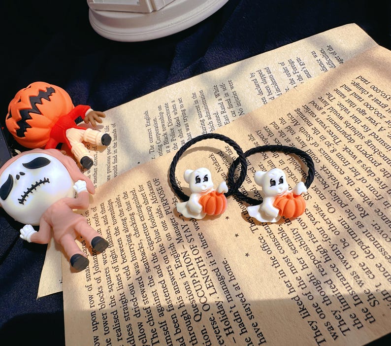 Pode incluir: Duas presilhas de cabelo com tema de Halloween com fantasmas brancos segurando ab&oacute;boras laranja. As faixas el&aacute;sticas pretas est&atilde;o em um fundo de papel vintage, com figuras de ab&oacute;bora e esqueleto. O tema &eacute; o Halloween.