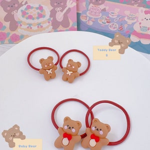 May include: Two sets of brown teddy bear hair ties with red elastic bands. One set has a white bow, the other has a red bow. The background includes illustrations of teddy bears and the words "birthday party".