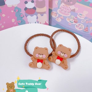 May include: Two brown hair ties with a cute teddy bear design. Each bear has a red bow, pink cheeks, and is attached to a brown, twisted elastic band. The background features a whimsical illustration with bears and floral elements.