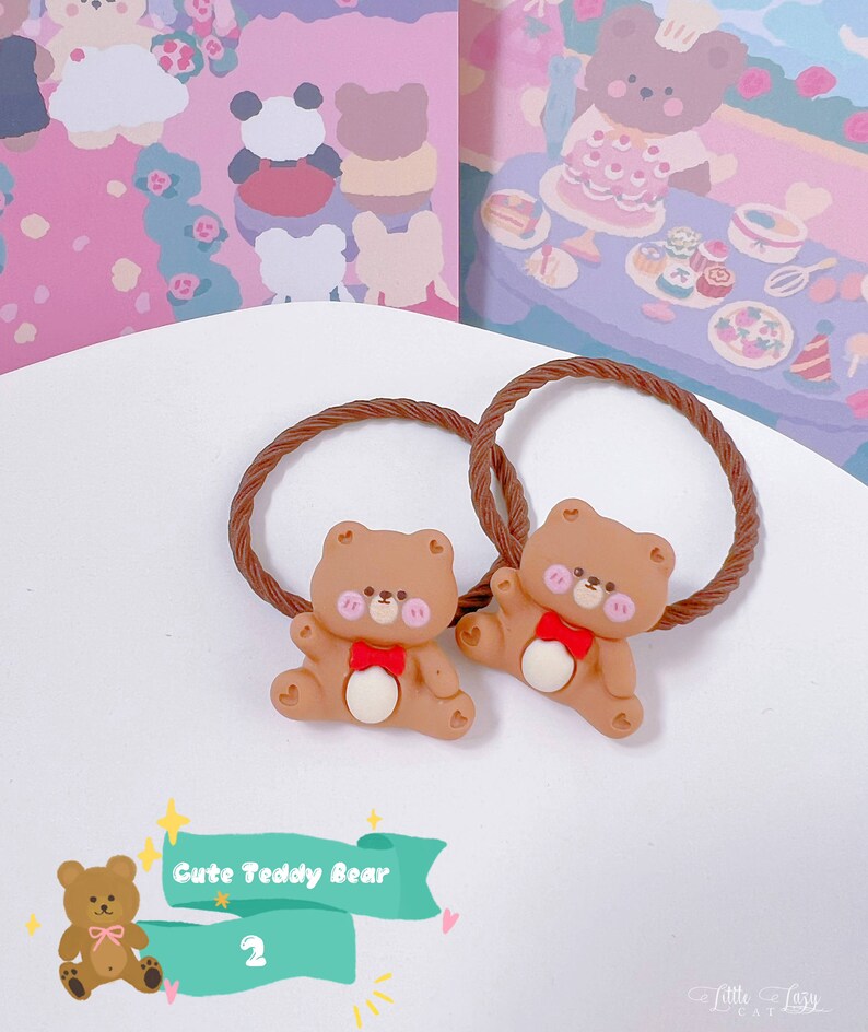 May include: Two brown hair ties with a cute teddy bear design. Each bear has a red bow, pink cheeks, and is attached to a brown, twisted elastic band. The background features a whimsical illustration with bears and floral elements.