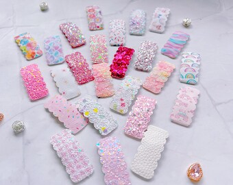 May include: A collection of decorative hair clips in various colors and patterns. The clips feature glitter, floral, and patterned designs, including rainbows, sprinkles, and a mermaid scale pattern. The clips are arranged on a white surface.