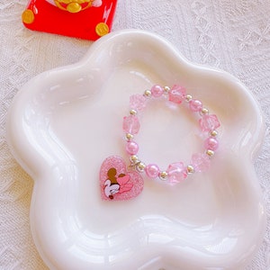May include: A pink beaded bracelet with a heart-shaped charm featuring a cartoon character. The bracelet has pink beads, clear square beads, and silver accents. The charm is glittery pink with a cartoon character design. The bracelet is displayed on a white, flower-shaped dish.