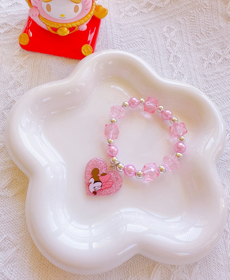 May include: A pink beaded bracelet with a heart-shaped charm featuring a cartoon character. The bracelet has pink beads, clear square beads, and silver accents. The charm is glittery pink with a cartoon character design. The bracelet is displayed on a white, flower-shaped dish.