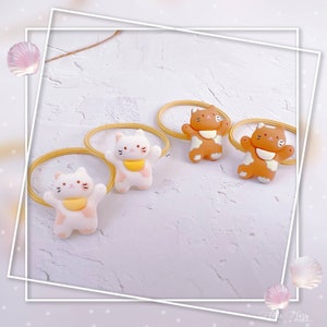 Puede incluir: Cuatro gomas para el pelo con forma de gato. Dos son blancas con manchas rosas y baberos amarillos, y dos son marrones con manchas blancas y baberos amarillos. Las gomas para el pelo son amarillas y est&aacute;n dispuestas sobre una superficie blanca texturizada.