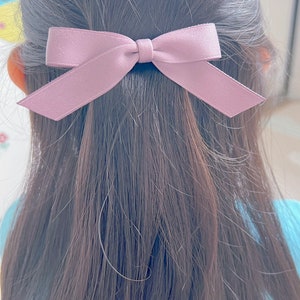 May include: A mauve bow hair clip is positioned in dark brown hair. The bow is made of a smooth, satin-like material. This simple hair accessory adds a touch of elegance.