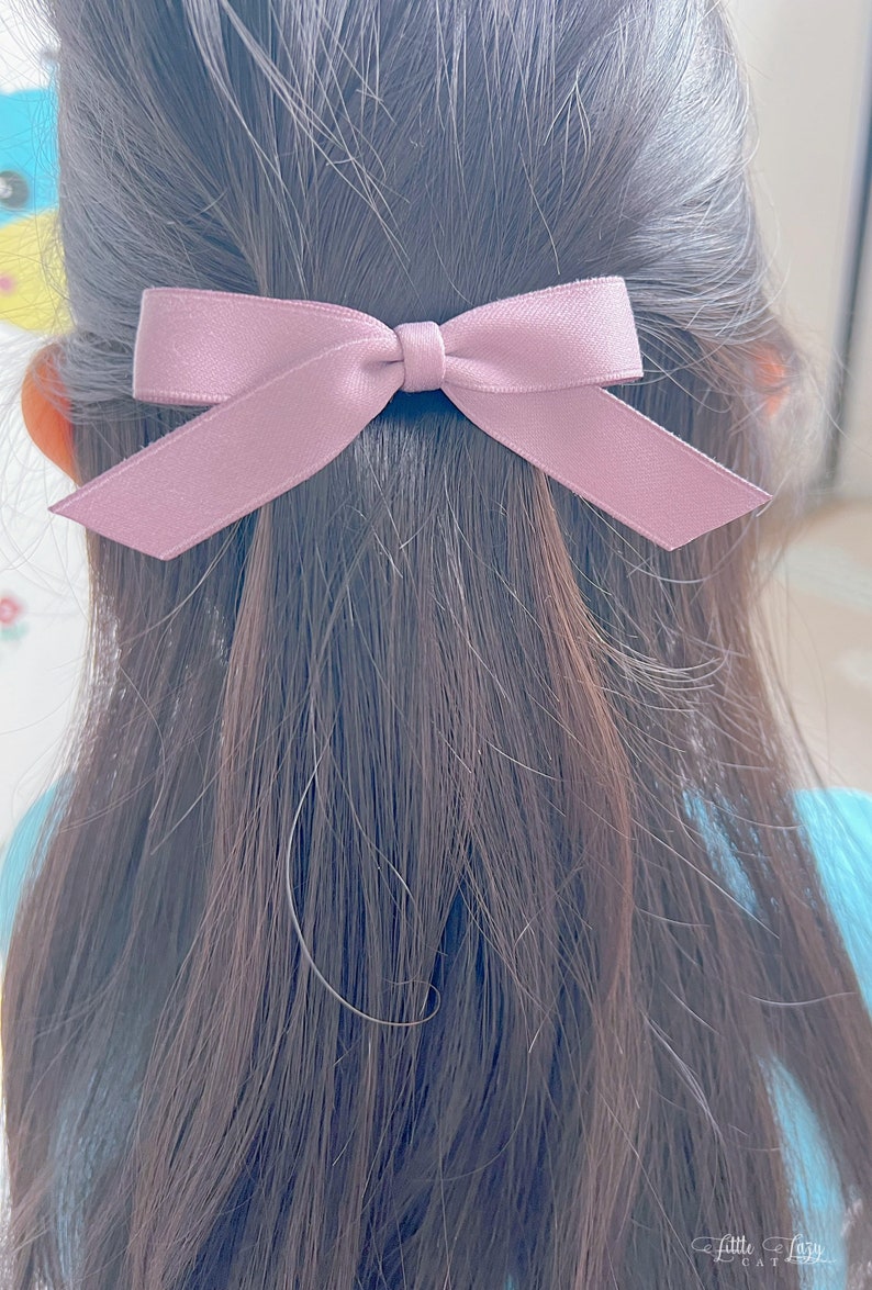 May include: A mauve bow hair clip is positioned in dark brown hair. The bow is made of a smooth, satin-like material. This simple hair accessory adds a touch of elegance.