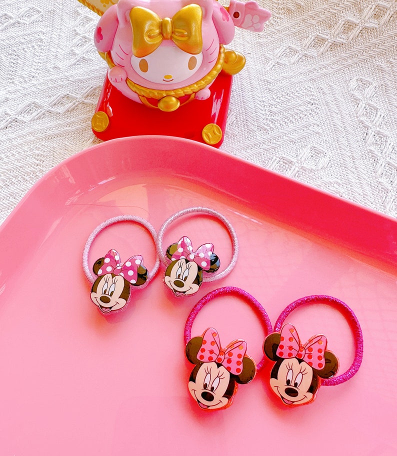 May include: Four pink Minnie Mouse hair ties with red polka dot bows and smiling faces are arranged on a pink tray. A pink and gold figurine is in the background.