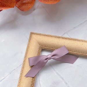May include: A small, mauve satin bow rests on the corner of a gold-coloured picture frame. The frame has a textured surface and a beaded border. Autumnal orange leaves are in the background, creating a seasonal aesthetic.