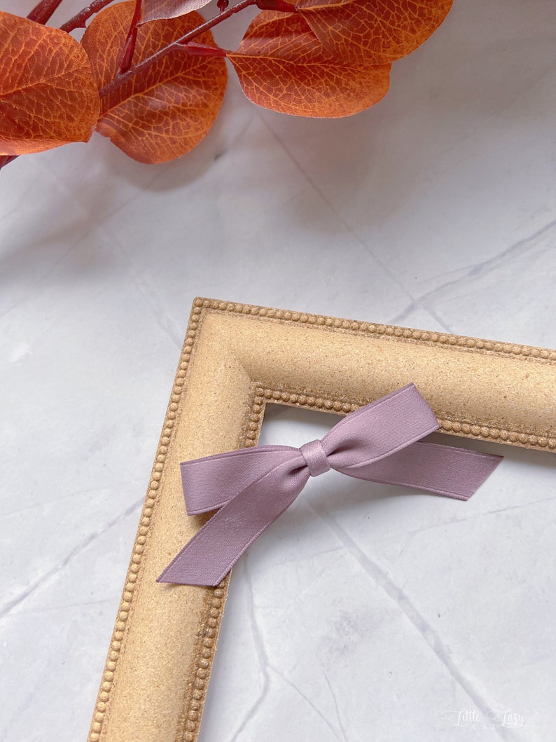 May include: A small, mauve satin bow rests on the corner of a gold-coloured picture frame. The frame has a textured surface and a beaded border. Autumnal orange leaves are in the background, creating a seasonal aesthetic.