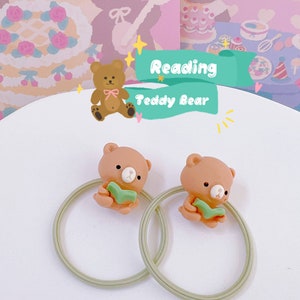 May include: Two hair ties featuring a light brown teddy bear reading a green book. The bears are attached to a light green elastic band. The background includes a cartoon illustration with the text "Reading Teddy Bear".