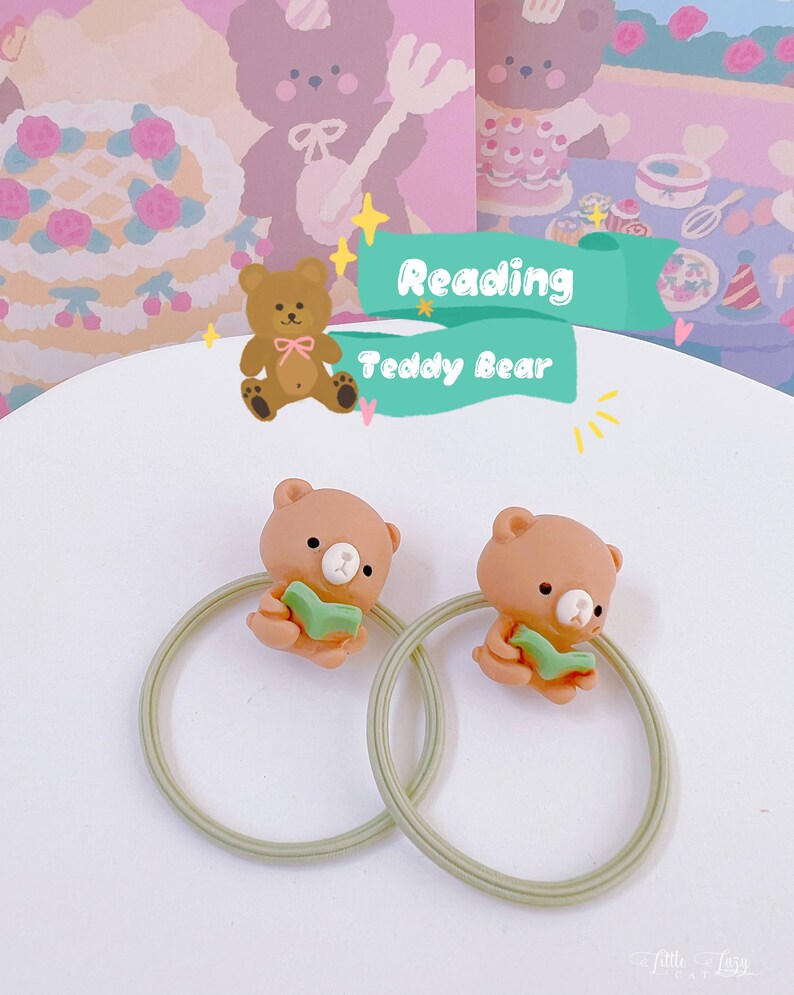 May include: Two hair ties featuring a light brown teddy bear reading a green book. The bears are attached to a light green elastic band. The background includes a cartoon illustration with the text "Reading Teddy Bear".
