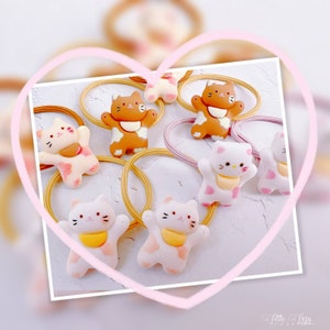 Puede incluir: Colecci&oacute;n de gomas para el pelo con forma de gato en blanco, marr&oacute;n y rosa. Las figuras de gato tienen baberos amarillos. Las gomas para el pelo est&aacute;n sobre una superficie blanca, enmarcadas por un coraz&oacute;n rosa. El texto "Little Lazy Cat" est&aacute; en la esquina inferior derecha.
