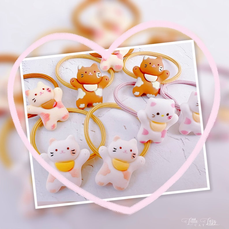 Puede incluir: Colecci&oacute;n de gomas para el pelo con forma de gato en blanco, marr&oacute;n y rosa. Las figuras de gato tienen baberos amarillos. Las gomas para el pelo est&aacute;n sobre una superficie blanca, enmarcadas por un coraz&oacute;n rosa. El texto "Little Lazy Cat" est&aacute; en la esquina inferior derecha.