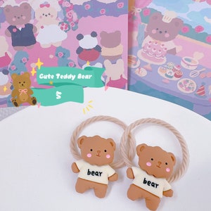 May include: Two brown teddy bear hair ties with beige rope-like bands. Each bear wears a white shirt with the word "bear" printed on it. The background includes a pink and purple illustration with teddy bear characters and the text "Cute Teddy Bear".